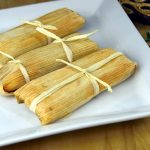 Three good tamales wrapped in corn husks tied with twine sit on a white square plate, resting on a wooden surface. A decorative item is partially visible in the corner, tempting you to eat tamales cold for a unique taste experience.