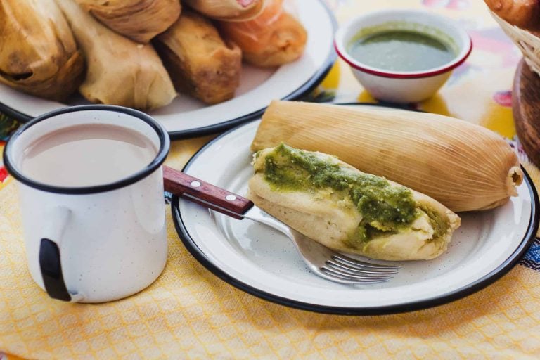 A plate with a tamale topped with green sauce, a fork, and an enameled mug of a hot beverage. In the background, more tamales await on a plate beside a small bowl of green sauce. The inviting scene rests on a yellow tablecloth, urging you to eat tamales in this cozy setting.