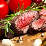 Slices of juicy, medium-rare venison steak garnished with rosemary, surrounded by garlic cloves, peppercorns, and ripe tomatoes on crumpled brown paper.