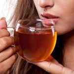 A person sips from a clear glass cup filled with amber-colored tea, savoring its warmth. Their lips are close to the cup's rim, with steam faintly visible against their face. The soothing tea is believed to ward off bloating, bringing moments of peace and comfort with each sip.