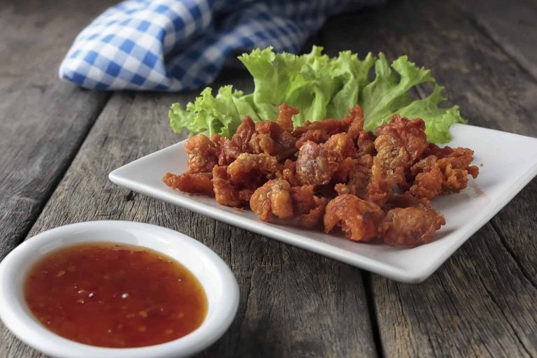 A square white plate filled with crispy fried pork and leftover chicken skin sits on a rustic wooden table. Fresh green lettuce is layered underneath. A small round dish beside it holds a vibrant red chili sauce. A blue and white checkered cloth is partially visible behind.