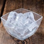 A square glass bowl filled with ice cubes sits on a wooden surface, inviting thoughts of eating ice—a curious habit that some say can even help alleviate bloating.