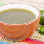 A bowl of green salsa in a red ceramic bowl sits on a colorful woven mat, infused with the tang of leftover jalapeño juice. Sliced jalapeños are visible in the background.