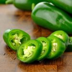 Sliced green jalapeño peppers, seeds exposed, are artfully arranged on a wooden surface, with whole peppers poised in the background, tempting you to eat them.