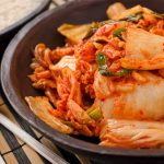 A bowl of spicy kimchi made with Napa cabbage and red chili flakes sits on a bamboo mat beside steaming white rice, releasing its distinct aroma. Silver chopsticks are placed beside the bowls, ready to delve into the fiery delight.