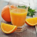 A glass of orange juice sits on a wooden table, flanked by two orange wedges and a whole orange. Fresh mint leaves enhance the vibrant and refreshing composition, offering a burst of zest that can help reduce bloating and gas naturally.