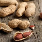 A close-up of peanuts on a wooden surface. Some are in their shells while others are cracked open, revealing reddish-brown seeds inside. The texture of the wood and peanut shells provides a rustic appearance, reminiscent of simpler times before worries about bloating or gas.