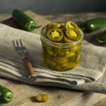 A glass jar filled with sliced pickled jalapeños sits on a rustic wooden table covered with a beige cloth. Whole jalapeños and a small fork are scattered around, alongside a hint of pickle juice, enhancing the rustic kitchen setting.
