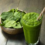 A tall glass filled with a green smoothie and a striped straw sits on a wooden table. Beside it, a bowl of fresh spinach leaves adds both nutrition and flair to the scene, making it perfect for those mindful of feeling bloated or gassy.