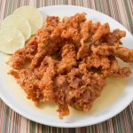 A white plate of crispy fried pork strips is served alongside three slices of lime, offering delicious reasons to indulge. The background features a striped pattern with various shades of red, green, and white.