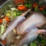 A large pot filled with a whole uncooked turkey immersed in a rich broth with chopped vegetables like carrots, leeks, and onions. Fresh herbs are sprinkled on top, creating an inviting soup base ready for cooking.