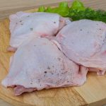 Three raw chicken thighs, with a few red spots sprinkled with pepper, rest on a wooden cutting board. Fresh green herbs, including lettuce and dill, are arranged beside the meat.