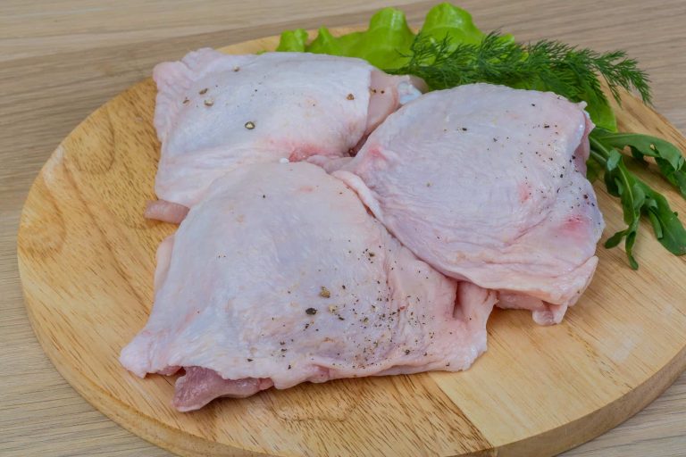 Three raw chicken thighs, with a few red spots sprinkled with pepper, rest on a wooden cutting board. Fresh green herbs, including lettuce and dill, are arranged beside the meat.