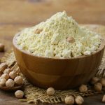 A wooden bowl filled with chickpea flour rests on a textured cloth surface. A few whole garbanzo beans are scattered around the bowl, with a wooden spoon holding more chickpeas nearby. The background is a rustic wooden table, perfect for crafting Paleo-friendly dishes.