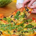 Close-up of a veggie pizza with fresh veggie toppings being lifted by two hands from a wooden table. The pizza is adorned with arugula, olives, bell peppers, and cheese. In the background, a salad bowl is slightly blurred.