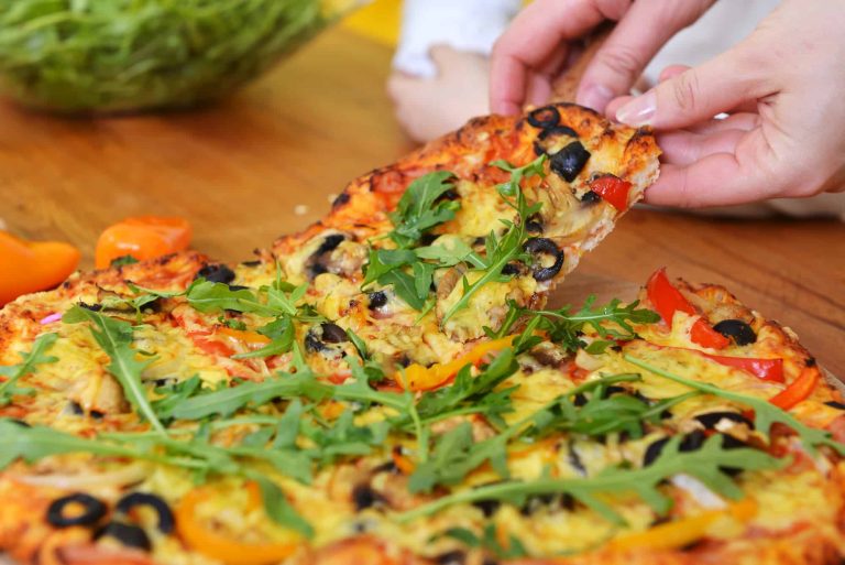 Close-up of a veggie pizza with fresh veggie toppings being lifted by two hands from a wooden table. The pizza is adorned with arugula, olives, bell peppers, and cheese. In the background, a salad bowl is slightly blurred.