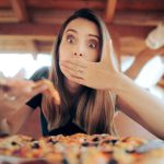 A surprised woman with long hair covers her mouth with one hand, savoring the taste of a slice of pizza with the other. She sits outdoors at a table, surrounded by a wooden roof structure, contemplating how different it is from her usual frozen pizzas.