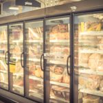 Blurred image of a grocery store's frozen food aisle with glass-door freezers displays organized freezer storage. Inside, various packaged frozen pizzas and other items are visible, each with a clearly marked expiry date. The atmosphere appears well-lit and efficient.