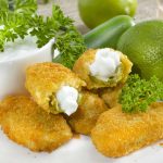 A plate of golden-brown jalapeño poppers with a creamy filling, garnished with fresh parsley, beckons. Accompanied by a bowl of zesty dip and a whole lime in the background, this dish offers irresistible sauce ideas for any occasion.