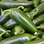 A close-up of a pile of fresh green jalapeño peppers, weighing several ounces. They are glossy and have a smooth texture, with stems visible on some of the peppers.