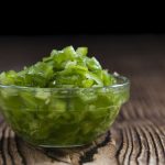 A small glass bowl filled with chopped green jalapeños is placed on a rustic wooden surface. The vibrant, diced peppers provide a spicy alternative to milder substitutes, creating a striking contrast against the dark background.