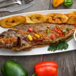 Grilled croaker fish served on a white rectangular plate, garnished with colorful chopped bell peppers, accompanied by fried plantains and a side of spicy dipping sauce. The dish is surrounded by fresh parsley, red onion, and bell peppers on a rustic wooden table.