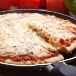 A freshly baked skillet pizza with a cheesy topping, seasoned with herbs. A slice is being lifted, showcasing gooey, melted cheese. In the background, fresh tomatoes and a green bell pepper rest on a wooden surface.