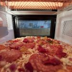 A pepperoni pizza topped with onions and peppers is inside a microwave, viewed from the front with the door open. As the oven's interior light illuminates the pizza during its cooking time, it heats perfectly to enjoy quickly without needing to cook frozen pizzas in advance.