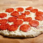 A store-bought frozen pepperoni pizza with a thin crust, topped with mozzarella cheese and slices of pepperoni, sits on a wooden surface. The pizza is uncooked, ready to be transferred from the fridge to the oven for baking.
