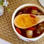 The image depicts a bowl of peach gum dessert, a traditional Asian delicacy. The dessert is served in a white bowl and includes a spoon showcasing its rich, jelly-like texture. Ingredients such as dried goji berries, longan, and snow fungus are visible, adding color and texture to the dish. The setup features a floral decoration, enhancing the aesthetic appeal of the scene.