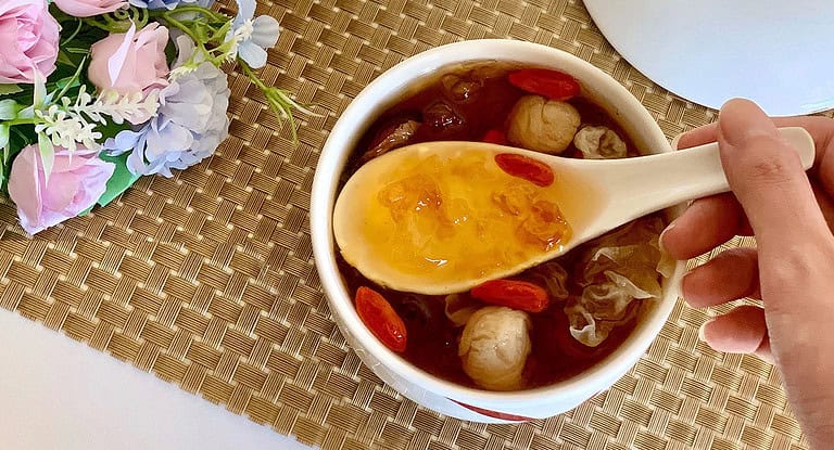 The image depicts a bowl of peach gum dessert, a traditional Asian delicacy. The dessert is served in a white bowl and includes a spoon showcasing its rich, jelly-like texture. Ingredients such as dried goji berries, longan, and snow fungus are visible, adding color and texture to the dish. The setup features a floral decoration, enhancing the aesthetic appeal of the scene.