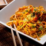 A white square plate filled with shrimp and vermicelli noodles, garnished with sesame seeds and fresh herbs, sits on a woven mat. Chopsticks rest beside the plate, while a small dish of sauce adds an extra layer of flavor in the background.