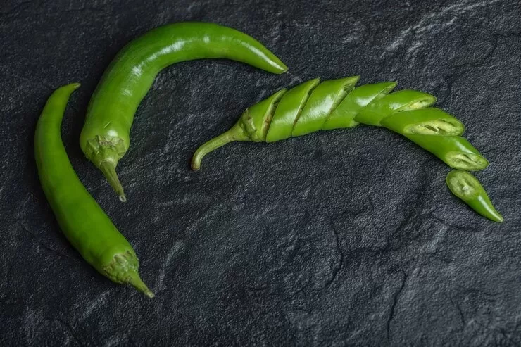 Three fresh jalapenos rest on a dark slate surface, with one pepper sliced into pieces.