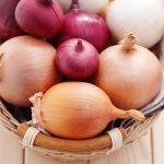 A woven basket filled with the best types of onions rests on a light wooden surface. The assortment includes red, yellow, and white onions, ideal for crafting fresh Pico de Gallo. Their papery skins add a mix of colors and textures, while a folded cloth sits beneath this vibrant array.