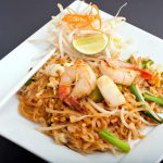 A plate of Pad Thai featuring vermicelli noodles, garnished with shrimp, bean sprouts, chopped peanuts, and lime. White chopsticks rest on the edge of the square plate, offering a delightful gluten-free option.