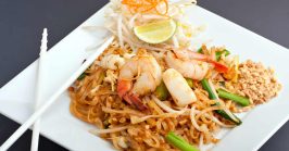 Seafood pad Thai dish and Thai fried rice noodles on a square white plate with chopsticks and grated carrot garnish.