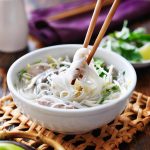 A bowl of pho with perfectly cooked noodles, meat, and herbs is being lifted by chopsticks. The bowl rests on a woven mat beside a dish of fresh herbs and lime, with a rich purple cloth in the background.