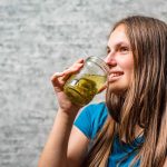 A person with long brown hair is smiling while holding a jar of pickles close to their face. They are wearing a blue shirt and standing against a textured gray background, perhaps inspired by the tangy allure of jalapeno juice.
