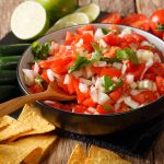 A bowl of fresh Pico de Gallo with diced tomatoes, onions, and cilantro sits on a wooden table surrounded by lime slices, whole tomatoes, jalapeños, and tortilla chips. A wooden spoon rests in the bowl, ready to scoop up the vibrant flavors and add a touch of heat.