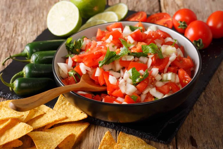 A bowl of fresh Pico de Gallo with diced tomatoes, onions, and cilantro sits on a wooden table surrounded by lime slices, whole tomatoes, jalapeños, and tortilla chips. A wooden spoon rests in the bowl, ready to scoop up the vibrant flavors and add a touch of heat.