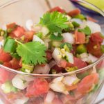 Close-up of a glass bowl filled with fresh Pico de Gallo, featuring diced tomatoes, onions, jalapeños, and cilantro leaves. The vibrant colors and textures highlight the authentic freshness of this USA-inspired salsa. Lime wedges are in the background.