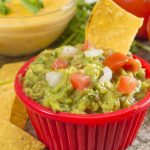 A red bowl brimming with guacamole, crowned with diced onions and tomatoes, is surrounded by crispy tortilla chips—perfect for enjoying leftovers or to serve pico de gallo. In the background, a bowl of cheese dip and two ripe tomatoes complete the inviting scene.