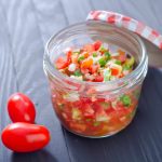 A clear jar with a checkered lid is packed with colorful diced vegetables, preserved for freshness, including tomatoes, onions, and herbs. Two cherry tomatoes rest beside the jar on a dark wooden surface.