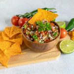 A wooden bowl of fresh pico de gallo garnished with parsley is surrounded by tortilla chips on a wooden board. Nearby are tomatoes, a lime cut in half, and green chili peppers, offering vibrant alternative ingredients on a light gray surface.
