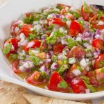 A bowl of homemade salsa with diced tomatoes, onions, cilantro, and orange bits is served with a silver spoon. This refreshing Pico de Gallo is accompanied by crunchy tortilla chips.