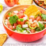 A red bowl filled with fresh, hot and spicy salsa features chopped tomatoes, onions, cilantro, and jalapeño slices. A tortilla chip dips into the mix resembling Pico de Gallo. Lime wedges and more chips are scattered around on a white wooden surface.