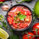 A vibrant bowl of Pico de Gallo, crafted with diced tomatoes, lime, and cilantro. Encircling the bowl are whole tomatoes, lime wedges, red and green chili peppers, garlic cloves, and parsley. For a fix to a watery mix, ensure your tomatoes are properly drained before dicing.