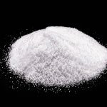 A mound of fine white salt sits against a black background, its granules scattered around the base. The surface is uneven and textured, much like potassium lactate crystals. While visually compelling, it's important to consider safety and risks before using substances like these in your diet.