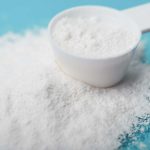 A white measuring scoop overflows with fine, white powder, spilling onto a bright blue surface. The soft texture of what could be potassium lactate is slightly dispersed around the scoop, standing out against the vibrant background.