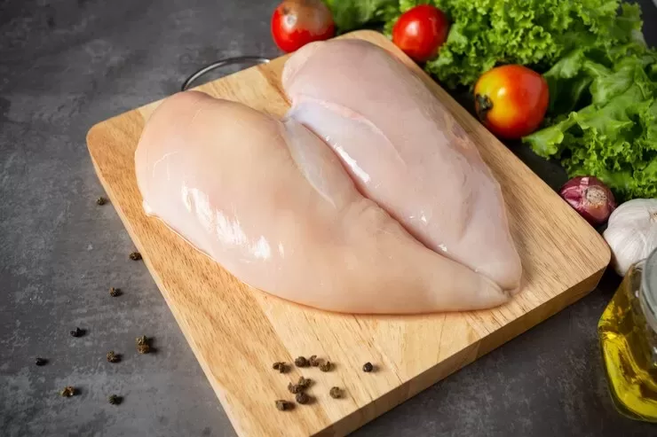 Two raw chicken breasts with subtle red spots rest on a wooden cutting board, surrounded by black peppercorns, ripe tomatoes, leafy green lettuce, and cloves of garlic. An olive oil bottle is visible on the side against a dark countertop background.